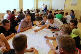 Grupa osób pochylająca się nad stołem, na którym rozłożone są mapy i dokumenty