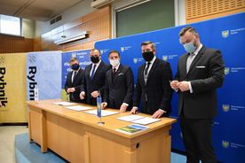 Pięciu mężczyzn w ciemnych garniturach i maseczkach na twarzy w trakcie konferencji prasowej. W tle banery reklamowe miasta Rybnika i subregionu zachodniego. 