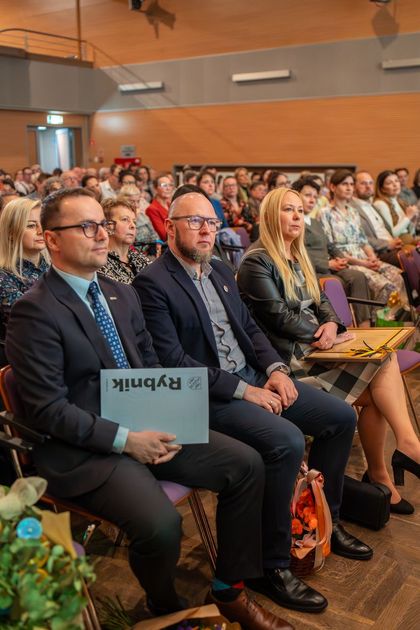 Grupa osób siedzi na widowni. Na pierwszym planie widać trzy osoby. 