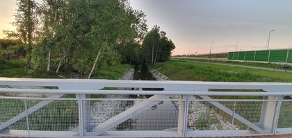 Balustrada mostu na rzece. Po obu stronach zieleń. Po prawej stronie ekrany akustyczne i barierki przy drodze szybkiego ruchu. 