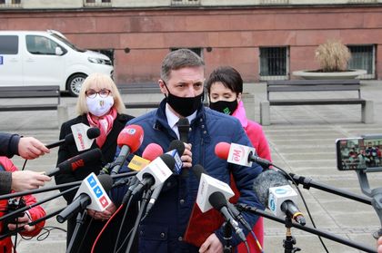 Briefing prasowy - mężczyzna i dwie kobiety stoją naprzeciwko ustawionych mikrofonów różnych redakcji radiowych i telewizyjnych 