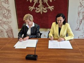 Zdjęcie przedstawia dwie kobiety siedzące przy biurku i podpisujące dokumenty.