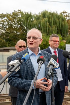 Zdjęcie przedstawia mężczyznę mówiącego do mikrofonu. W tle widać dwóch mężczyzn.