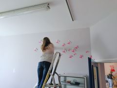Kobieta na drabinie przykleja na białej ścianie różowe motylki. 