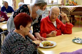 Starsze osoby spożywają posiłek przy stolikach na dużej, jasnej sali. na pierwszym planie kobieta i mężczyzna przy stole, obok kobieta w maseczce podaje jedzenie. 