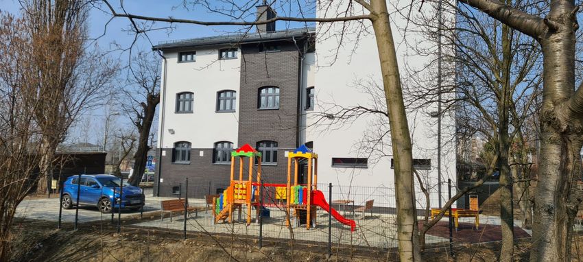 Biały, dwupiętorowy budynek otoczony płotem, za płotem plac zabaw