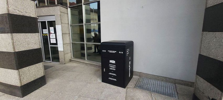 Czarna, metalowa wrzutnia na dokumenty urzędowe ustawiona przed budynkiem