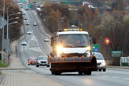 Ulice miasta, na nich pługopiaskarka i pracownicy służb komunalnych z szuflami do odśnieżania.