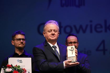 Mężczyzna w ciemnym garniturze ze statuetką w prawej ręce. W tle dwóch mężczyzn w okularach.