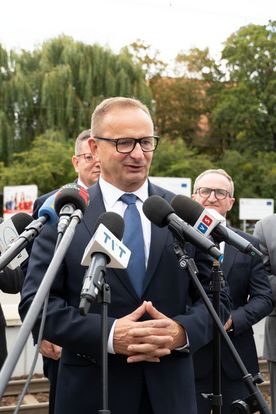 Zdjęcie przedstawia mężczyznę, który mówi do mikrofonu. W tle widać dwóch mężczyzn.