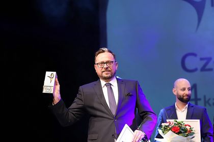 Mężczyzna w ciemnym garniturze ze statuetką w prawej ręce. W tle mężczyzna. 