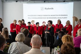 Grupa kobiet stoi przy mikrofonie. W tle grupa siedzących ludzi.