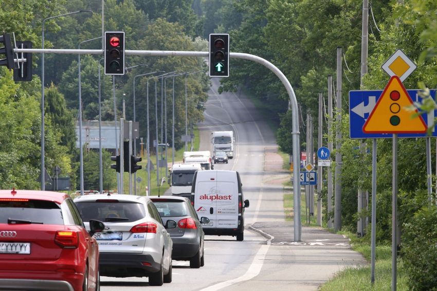Droga dwukierunkowa otoczona lasem, jadące nią samochody. Nad drogą sygnalizacja świetlna