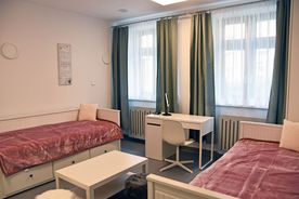 Pokój sypialny, jasne wnętrza, dwa łóżka, duże okna z zasłonami, na środku pokoju biały stolik.