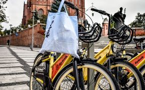 Żółty rower miejski, stojący na placu przed bazyliką. Na kierownicy wisi błękitna torba z napisem Rybnik.