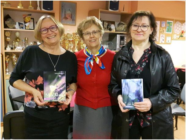 Trzy kobiety w okularach. Pani w środku w czerwonym swetrze, panie na zewnątrz ubrane w ciemne kolory, obie trzymają w rękach książkę. 