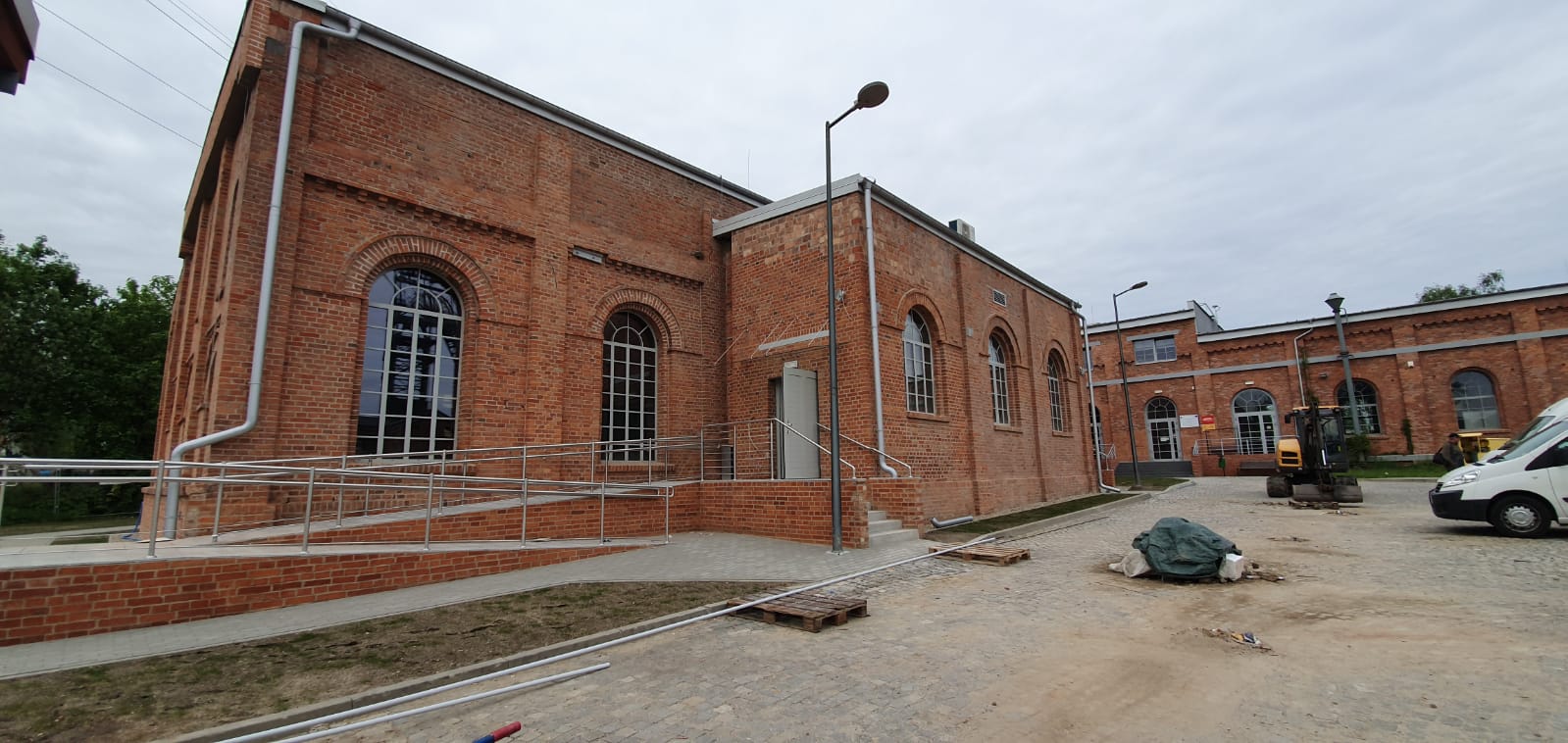 Ignacy 05 2020 0 Budynek z czerwonej cegły z podjazdem dla osób niepełnosprawnych. Przed budynkiem plac, na którym są: koparka, samochód i śmieci budowlane.