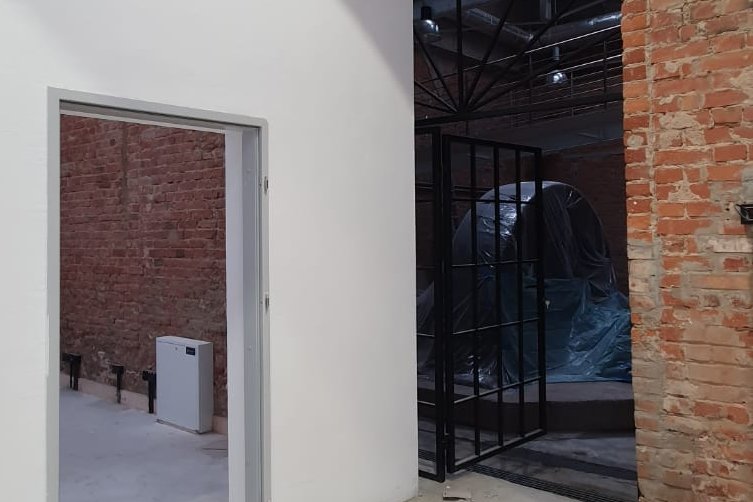 Ignacy 05 2020 14 Industrialne wnętrze, ściany z czerwonej cegły, stalowe konstrukcje. Po lewej stronie biała otynkowana ściana z otwartymi drzwiami.