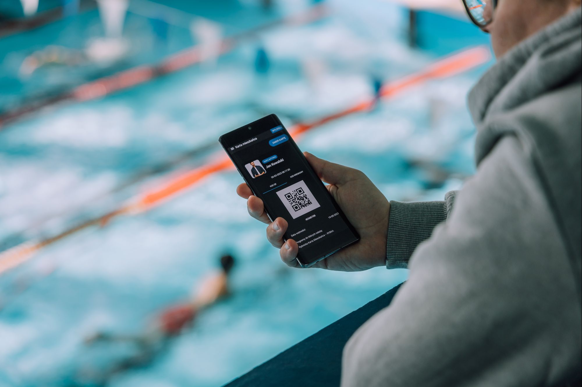 Osoba trzymająca spartfon z otwart aplikacja karty "Rybka" w tle pływalnia